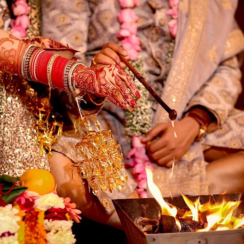 A couple participates in a traditional wedding ceremony, with decorated hands holding items over a fire and surrounded by floral arrangements.