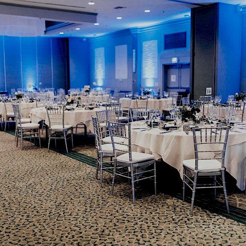 A banquet hall is shown with tables set for an event. It features blue lighting, round tables with white tablecloths, and metal chairs.