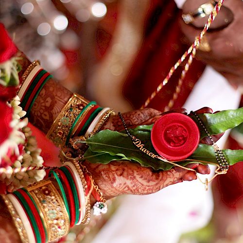 Two hands adorned with traditional henna and jewelry hold a small ceremonial arrangement with leaves and a red item, part of a cultural ceremony.