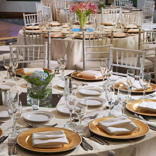 The image shows an elegantly set dining area with round tables, white chairs, gold-rimmed plates, and floral centerpieces in a well-lit room.