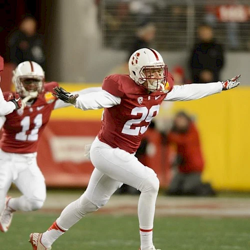 Two football players in red and white uniforms are celebrating on the field. One player has arms outstretched, and the other is in the background.