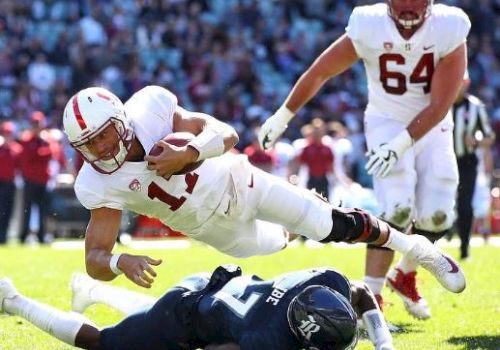 A football player in white dives over an opponent in blue, holding the ball, with a teammate behind him and a crowd in the background.