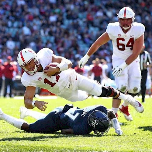 A football player in white dives over an opponent in blue, holding the ball, with a teammate behind him and a crowd in the background.
