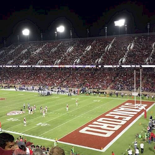 A nighttime football game is happening in a stadium, with players on the field and spectators filling the stands, under bright stadium lights.