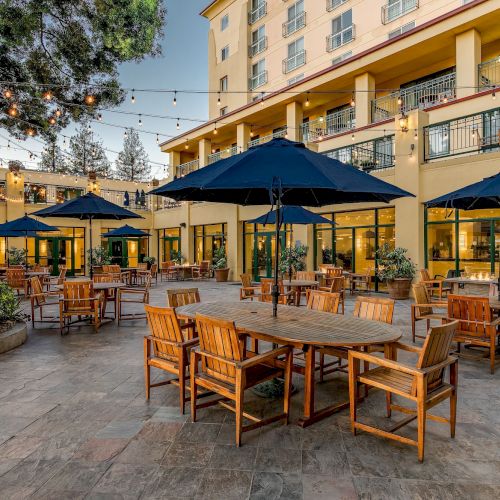 An outdoor patio area with wooden tables and chairs, large blue umbrellas, string lights, and a view of a multi-story building.