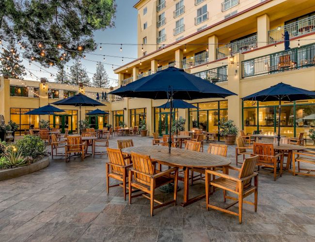 An outdoor patio area with wooden tables and chairs, large blue umbrellas, string lights, and a view of a multi-story building.