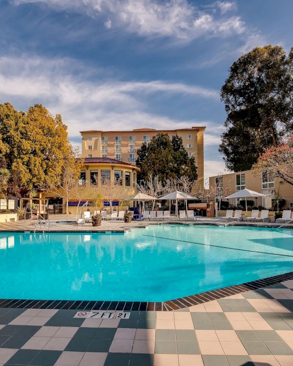 A serene outdoor pool area with lounge chairs, umbrellas, and nearby buildings surrounded by trees under a partly cloudy sky.