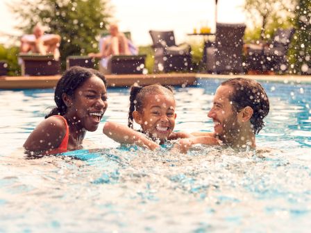 Three people are happily splashing in a swimming pool, while others relax in the background on lounge chairs, enjoying a sunny day.