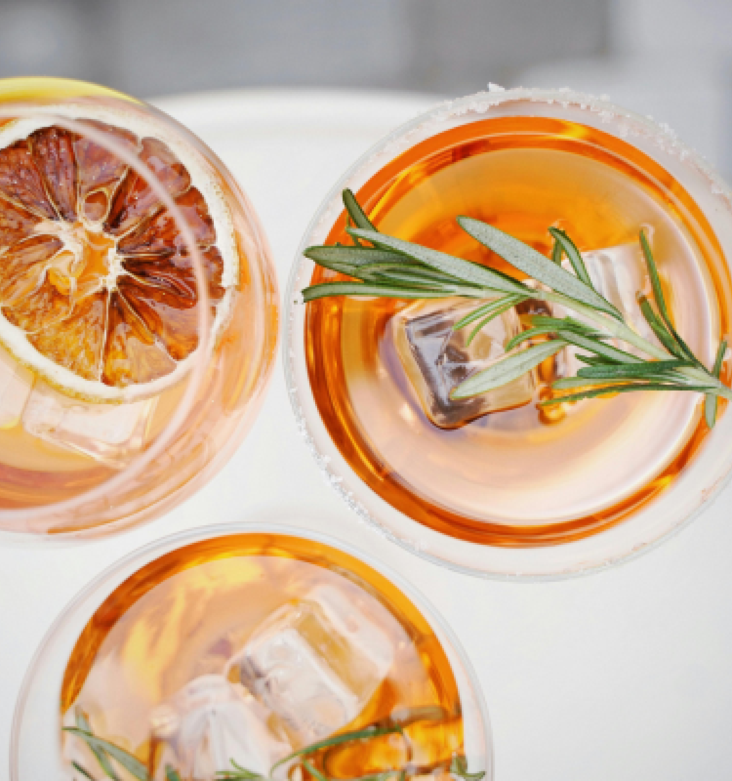 Three glasses with iced drinks, garnished with rosemary and a slice of dried citrus, viewed from above.