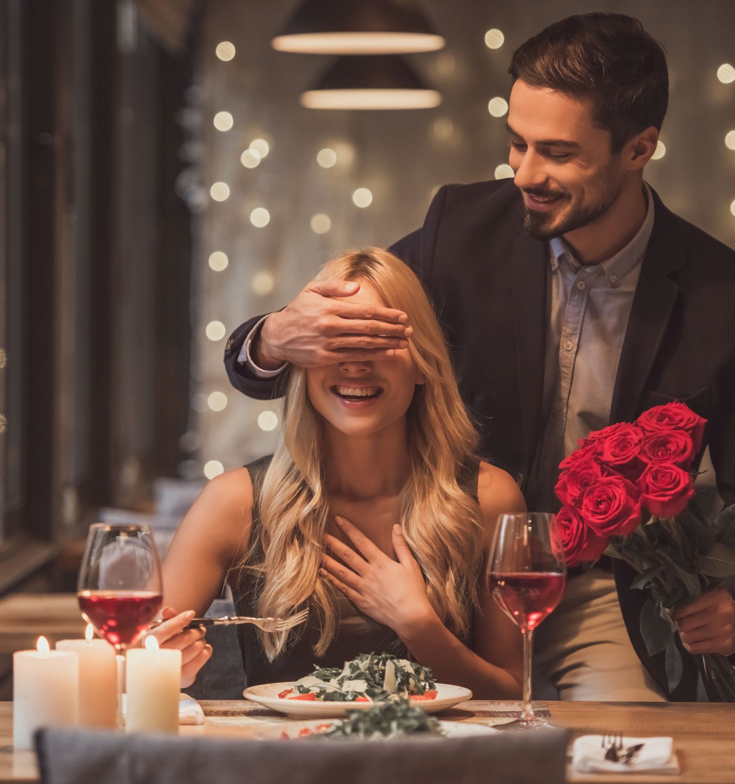 A man surprises a woman in a restaurant, covering her eyes and holding red roses. Candles and wine adorn the table.