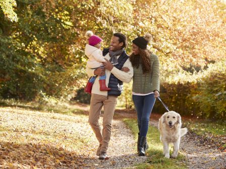 A family is enjoying a walk in a park with autumn foliage, carrying a baby and walking a dog on a leash.