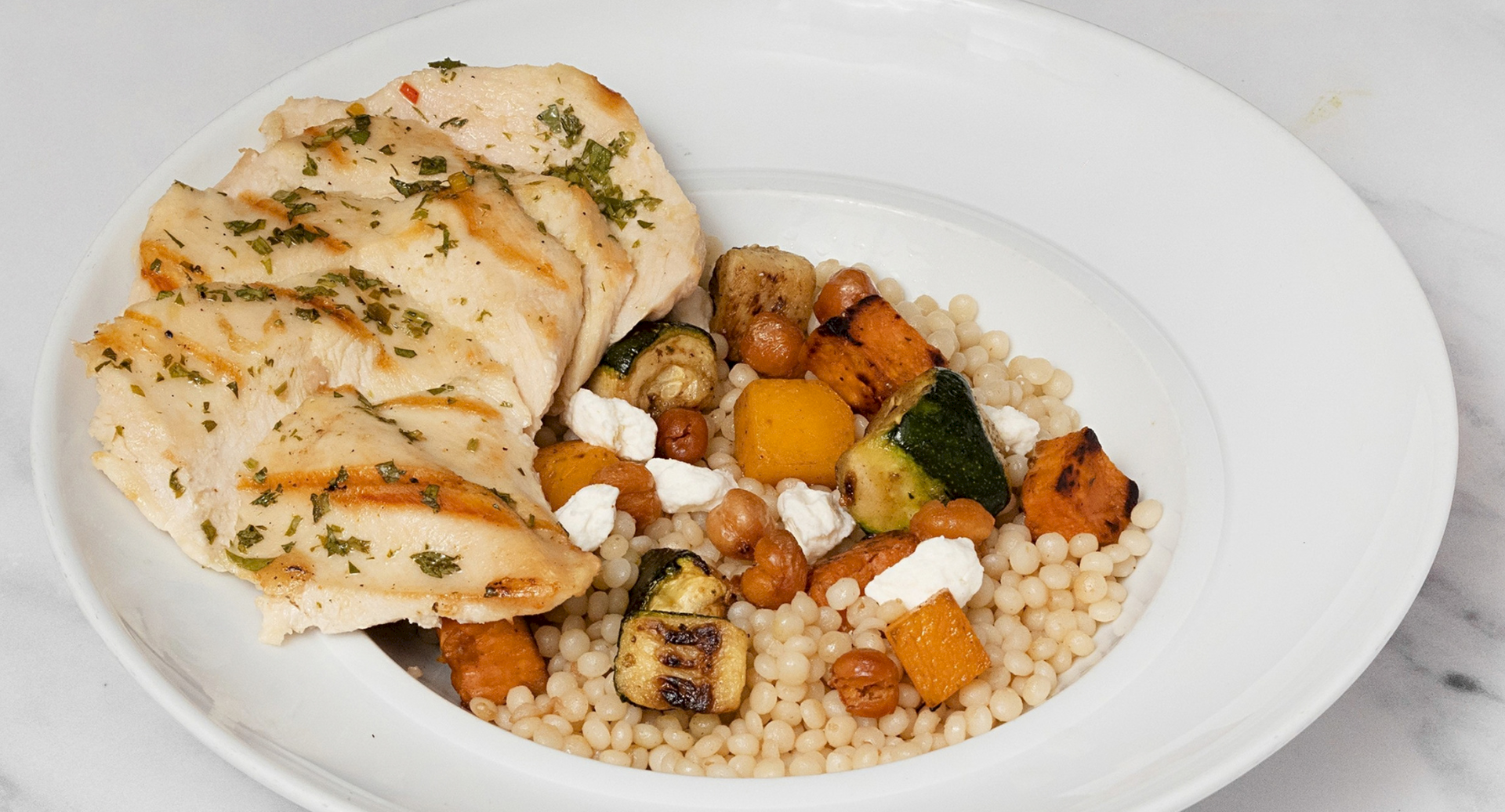 A plate of herb-roasted chicken over couscous with roasted vegetables and crumbled feta.