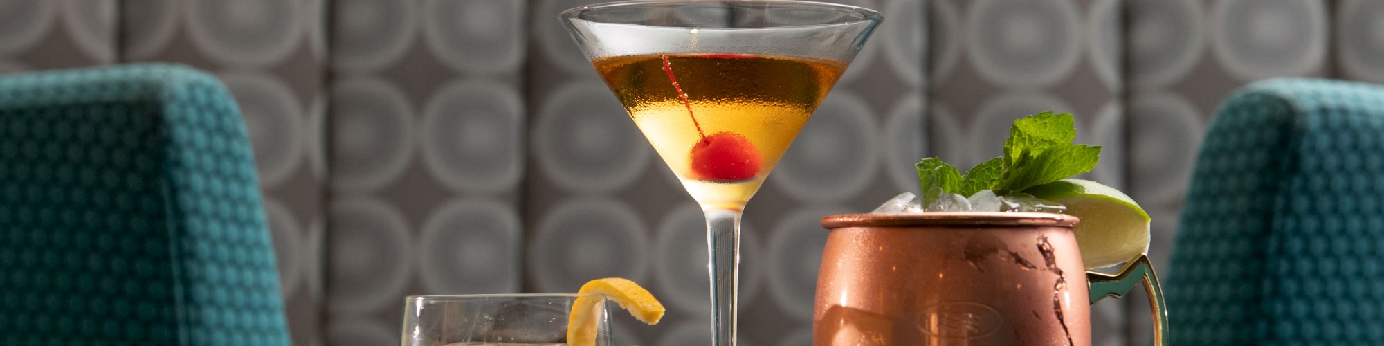 A trio of cocktails on a glass table: a short glass with lemon, a martini with an olive, and a copper mug with mint garnish&mdash;stylish drinks.