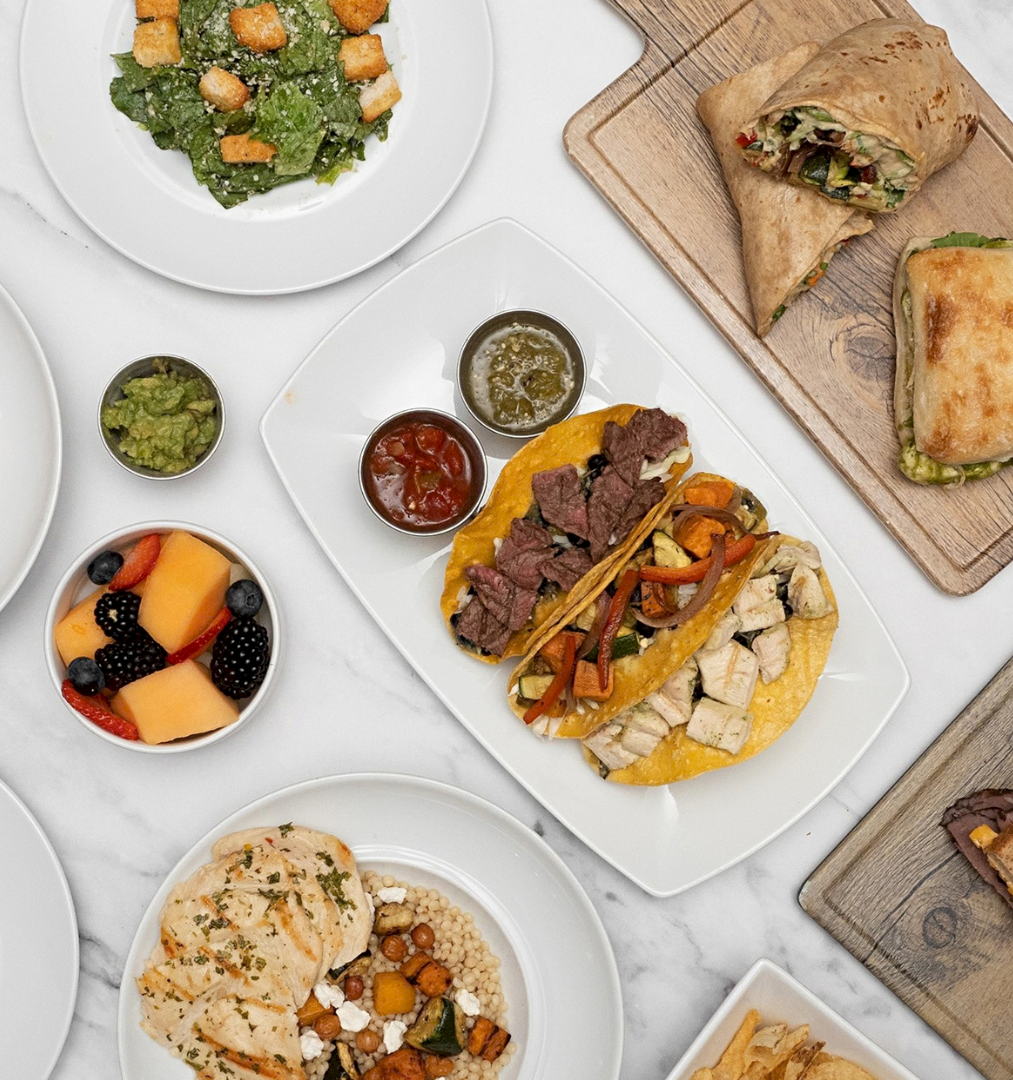 A spread of plated meals&mdash;meatballs and pasta, tacos/flatbread wraps, sandwiches, salad, and fruit&mdash;served on marble tables.