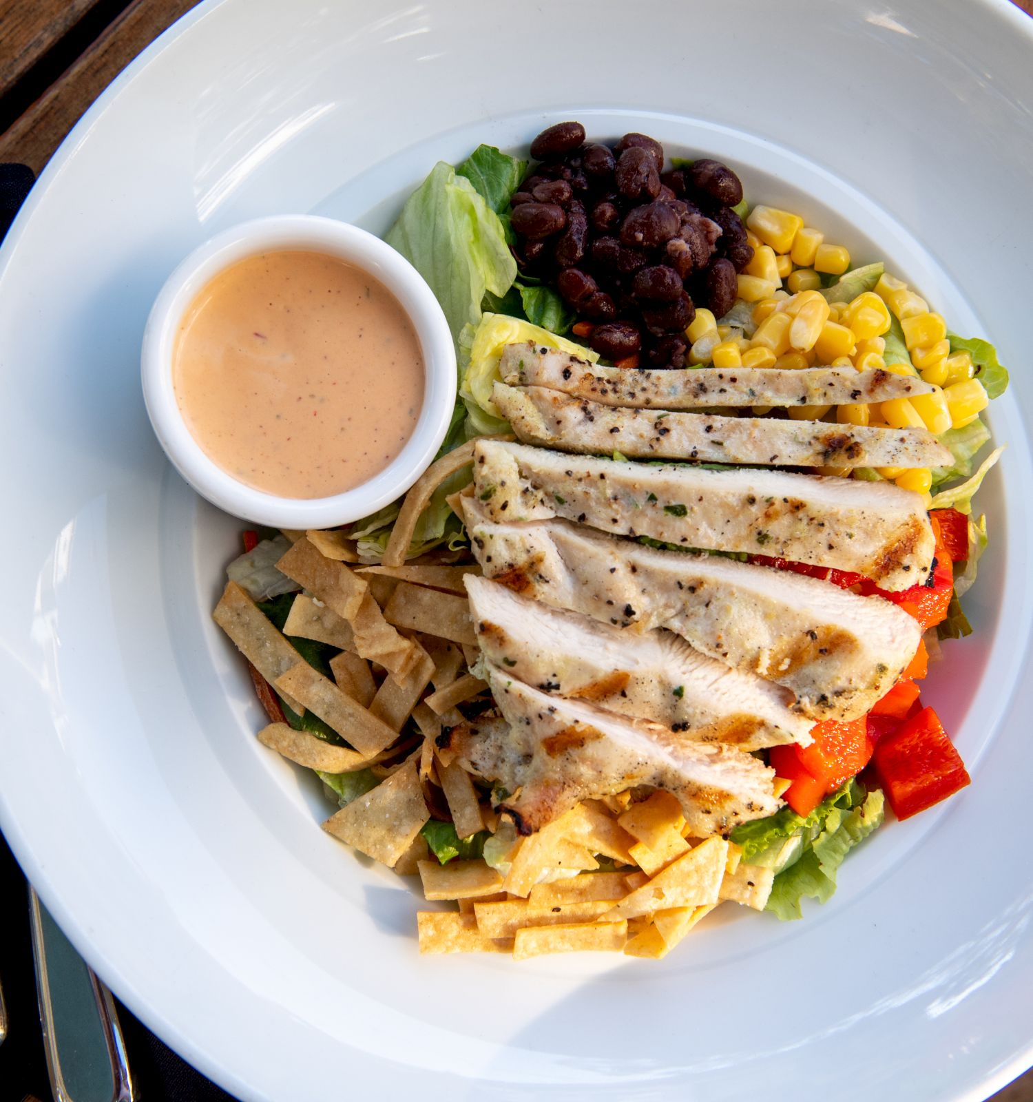 A salad bowl with grilled chicken slices, corn, black beans, tortilla chips, diced tomatoes, greens, a side of dressing, all on a white plate.