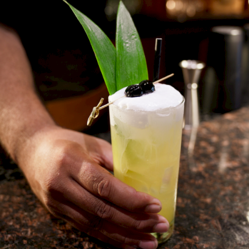 A bartender mixes a tall yellow-green cocktail with mint, served in a frosted glass, topped with a lime wedge.