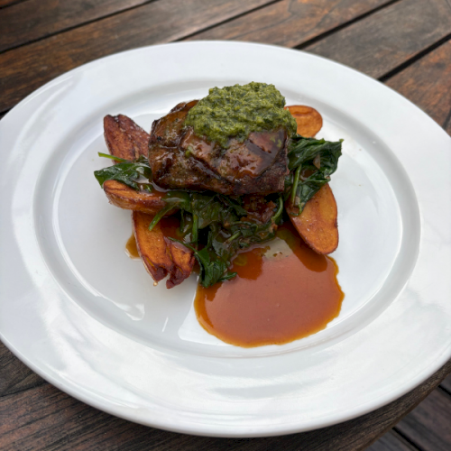 Grilled steak atop wilted greens with roasted carrots, a dollop of green herb sauce, and a glossy sauce drizzle on a white plate.