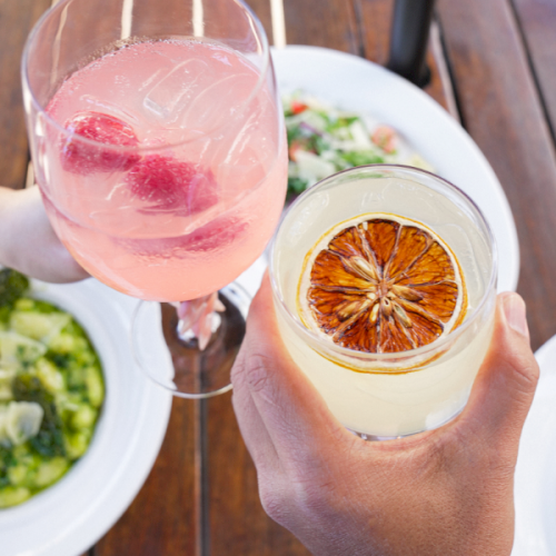 Two gloved hands hold drinks: a pink cocktail with a lime wheel and a light brown beverage topped with a citrus slice, with a green salad nearby.