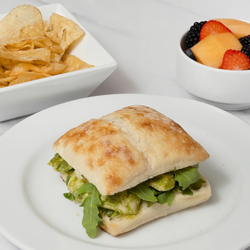 A turkey-style sandwich with greens, a side of potato chips, and a bowl of fresh fruit like strawberries, melon, and berries.