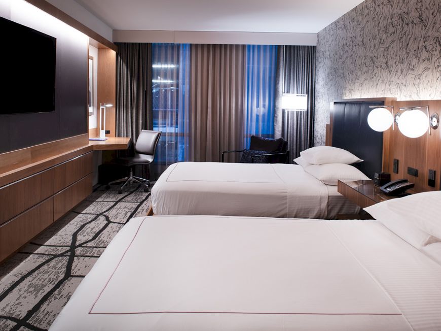 A modern hotel room features two neatly made beds, a wall-mounted TV, a desk with a chair, and warm lighting from lamps on nightstands.