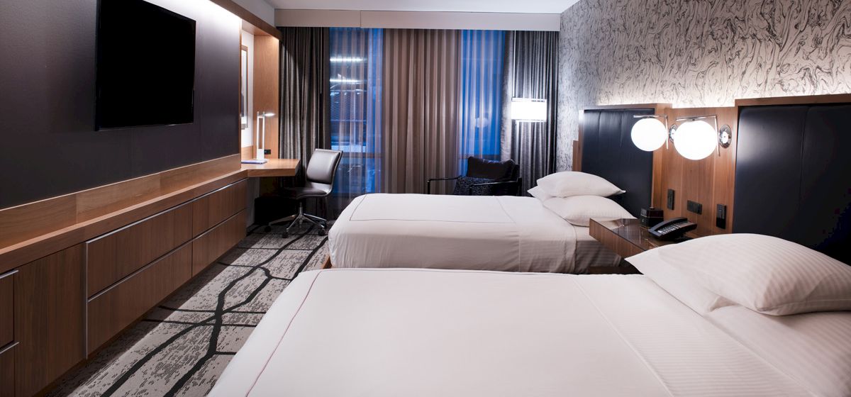 A modern hotel room features two neatly made beds, a wall-mounted TV, a desk with a chair, and warm lighting from lamps on nightstands.