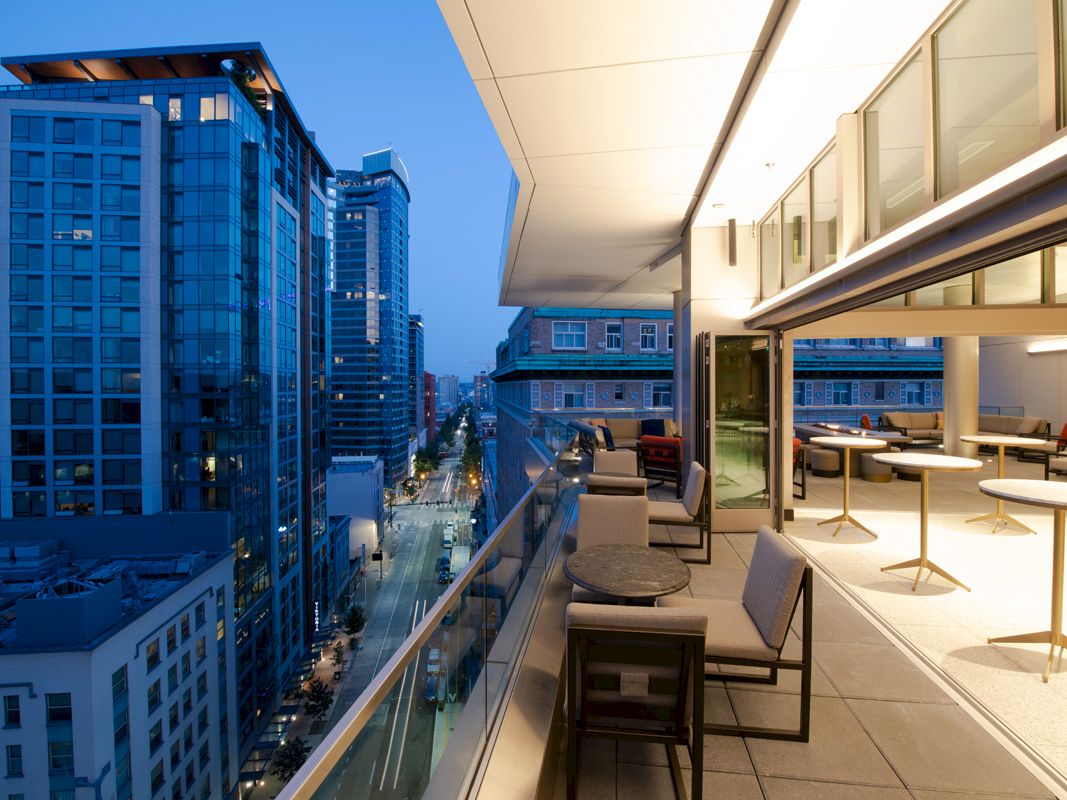 A modern cityscape at dusk featuring a sleek balcony with tables and chairs overlooking city buildings and a street below.