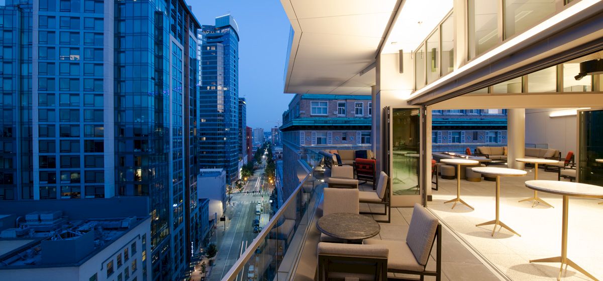 A modern cityscape at dusk featuring a sleek balcony with tables and chairs overlooking city buildings and a street below.