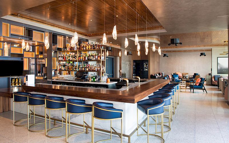 A modern bar area with a well-stocked shelf, a wooden counter, blue bar stools, pendant lighting, and seating areas in the background.