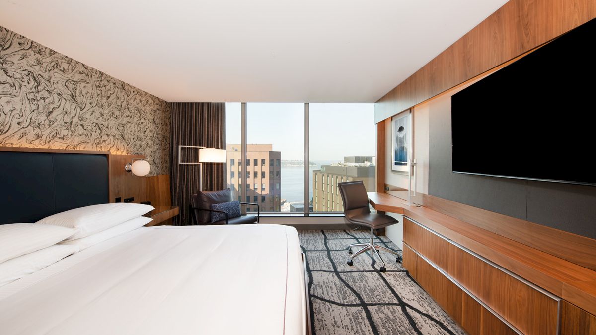 A modern hotel room with a large bed, wall-mounted TV, desk, chair, and a window offering a city view. Neutral tones and stylish decor.